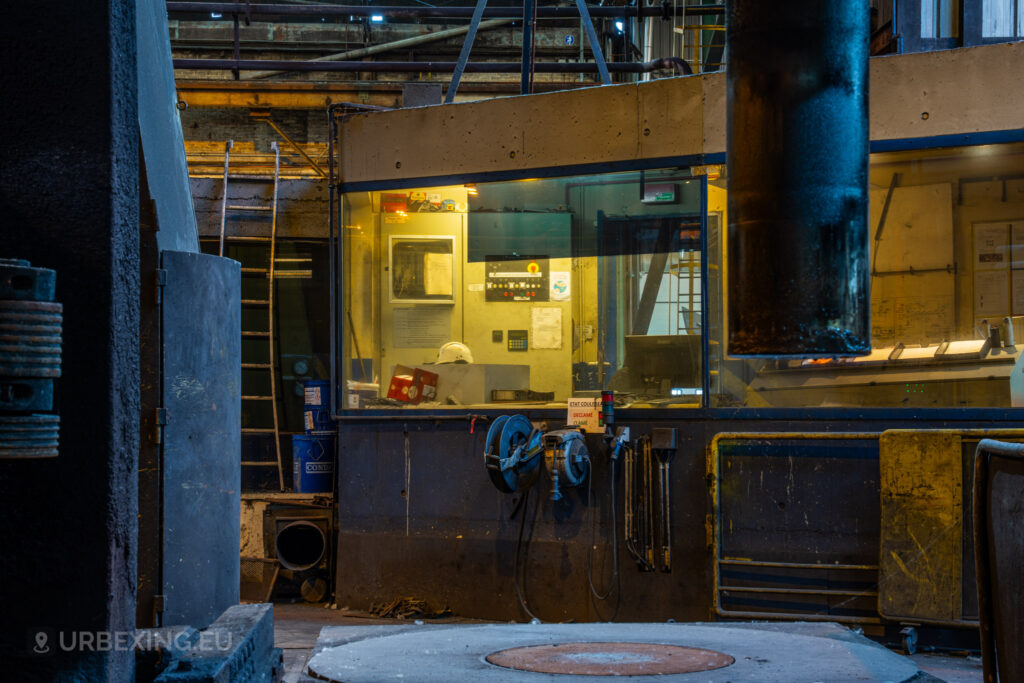 A dimly lit view of an operator cabin inside the abandoned Cockerill Forges & Ringmill factory. The cabin is illuminated from within, revealing scattered paperwork, a safety helmet, and control panels. Outside the cabin, industrial pipes, a ladder, and coiled hoses emphasize the once-busy nature of the workspace. The dark, heavy machinery in the foreground and the overall state of disrepair evoke the atmosphere of a factory that has been left untouched for years.