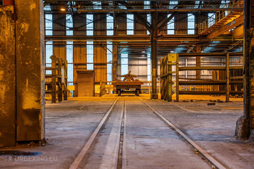 A view down an old rail track running through the Cockerill Forges & Ringmill factory, leading to a platform with a heavy cart holding industrial components. The scene is illuminated by dim, orange overhead lights that cast a warm glow across the space. Metal frames, barriers, and shelving units are visible along the sides, with a high ceiling supported by structural beams. The large windows in the background allow light to filter in, revealing the expansive, empty interior of the factory.