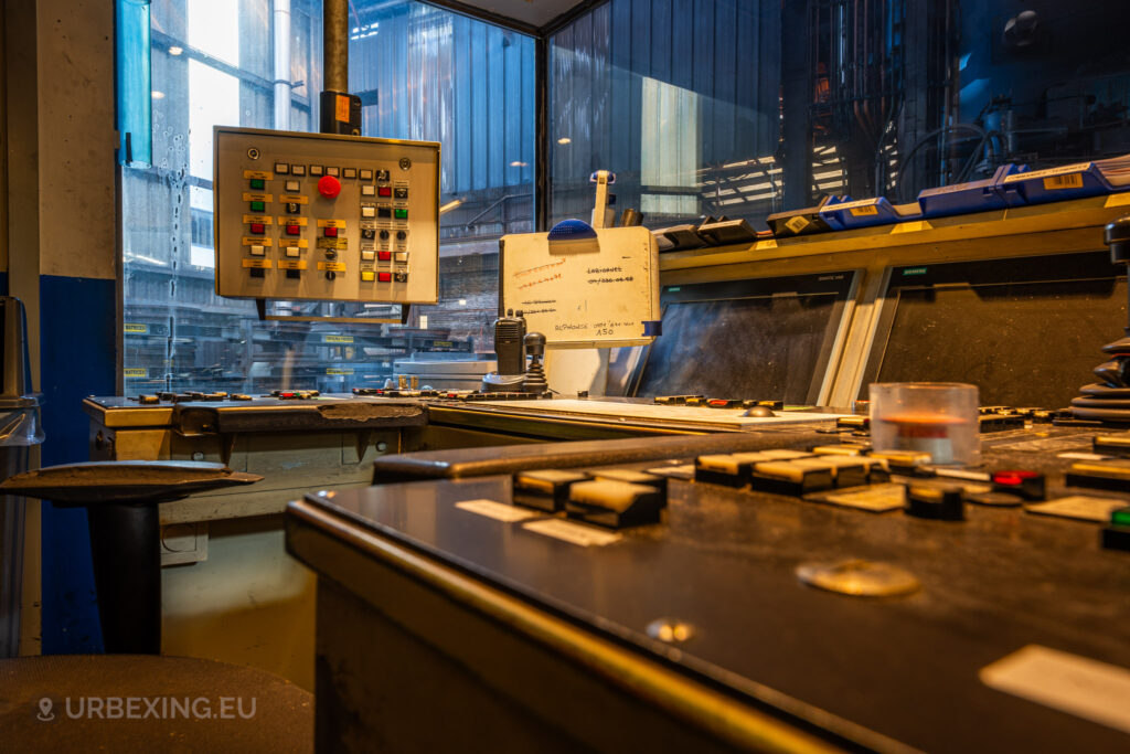 Close-up view of an abandoned control panel in the command room of the Halo Steelrings / Cockerill Forges & Ringmill factory. The image features a panel filled with various buttons, switches, and a large red emergency stop button. In the background, a dirty window provides a partial view of the abandoned factory floor, with industrial structures barely visible through the glass. Dust and grime cover the surfaces, hinting at long-term disuse of the equipment.