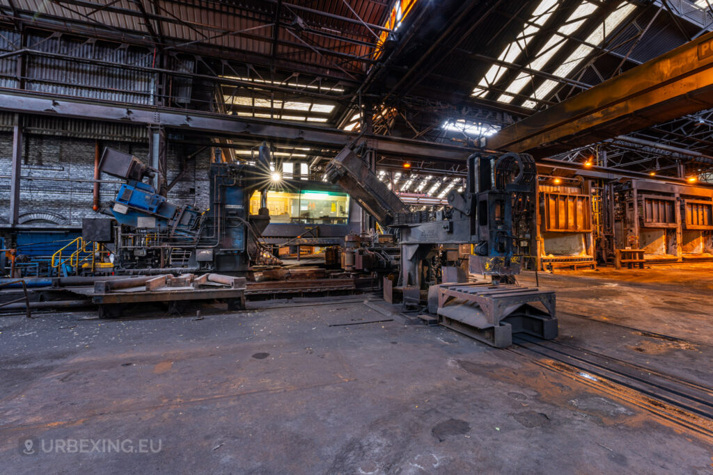 A view inside the abandoned Halo Steelrings / Cockerill Forges & Ringmill factory, showcasing a long row of rusted industrial machinery. The large, mechanical structures, beams, and rails on the ground highlight the scale of former metalworking operations. The spacious factory floor is marked with grime and wear, with a brightly lit control room in the background. Overhead, the steel trusses and skylights bring some natural light into the otherwise dim and deteriorating interior.