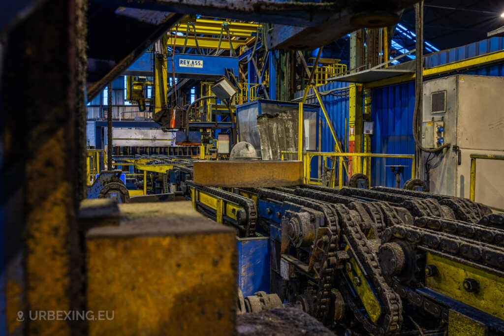 A close-up view of rusty conveyor chains and industrial machinery in the abandoned Cockerill Forges & Ringmill factory. The scene features a complex system of chains, gears, and overhead equipment, with machinery labeled ‘REV.ASS’ visible in the background. The blue and yellow components are heavily corroded, showing signs of long-term neglect. The various pipes, safety railings, and control panels emphasize the heavy-duty industrial environment of the steel production facility.