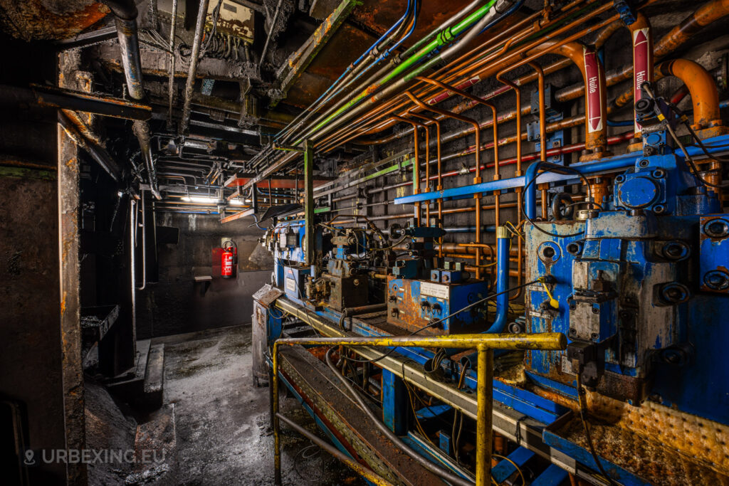 An interior view of a mechanical control room in the abandoned Cockerill Forges & Ringmill factory, showcasing a maze of corroded pipes and rusty machinery. The walls are lined with multicolored pipes, valves, and old hydraulic systems. The machinery is covered in grime, reflecting the long-term neglect of the facility. Dim lighting creates shadows across the scene, while a red fire extinguisher hangs on the wall, contrasting against the dark, worn surroundings of this industrial space.