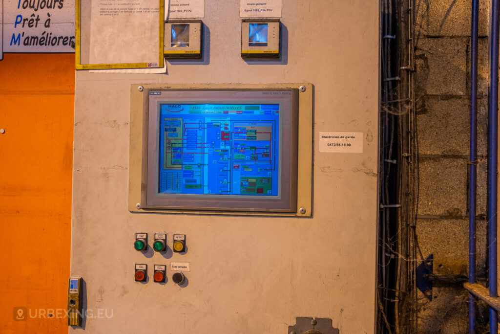 A close-up view of a touchscreen control panel in the abandoned Cockerill Forges & Ringmill factory. The Siemens SIMATIC Multi Panel display shows a complex system diagram, while below, there are colored buttons labeled ‘Test lampes’ and ‘Reset.’ Nearby, there is a note with a phone number labeled ‘Électricien de garde’ for emergency assistance. The area is surrounded by wiring and conduits, and the wall shows signs of wear and grime, indicating the age and neglect of the facility.