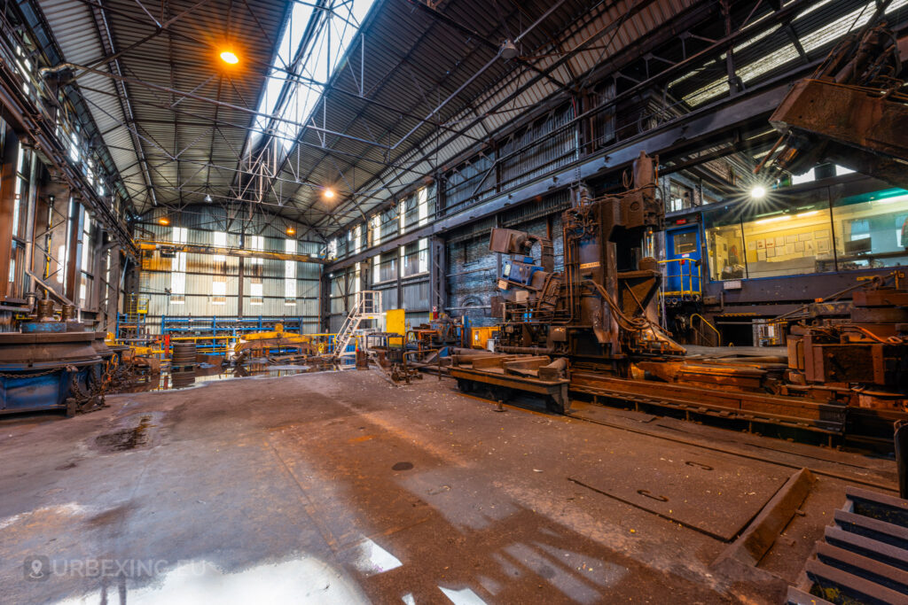 An expansive view of the workshop floor inside the abandoned Cockerill Forges & Ringmill factory, showing large industrial machines and a control room with illuminated windows. The workshop features a high ceiling supported by metal beams and skylights, allowing light to filter in. Heavy machinery lines the sides of the space, while scattered equipment, rail tracks, and puddles cover the dusty floor. The rust and decay indicate years of abandonment in this vast steel processing facility.