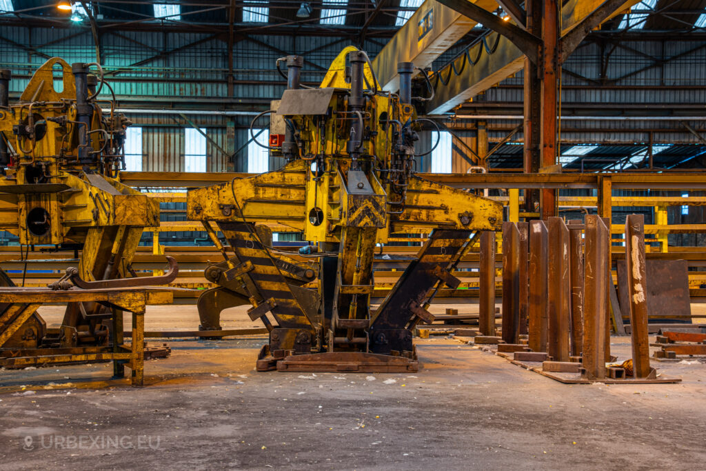 A close-up view of large, yellow industrial clamp machinery at the Cockerill Forges & Ringmill factory. The machinery, with hydraulic components and multiple moving parts, sits idle in the abandoned forge. Nearby are rows of metal beams, and the background reveals the high-ceilinged interior of the factory, with structural metal beams and overhead cranes. The yellow paint is worn, indicating extensive past use, and the overall scene conveys a sense of heavy industry left behind.