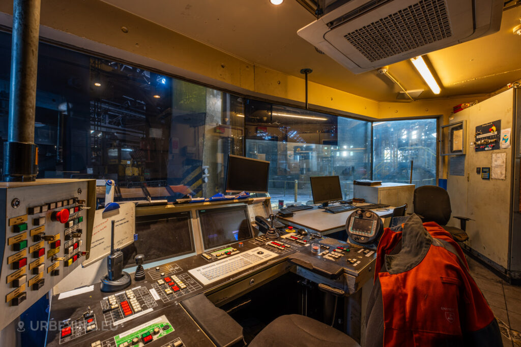 A detailed view of the control room at Halo Steelrings / Cockerill Forges & Ringmill, showcasing dusty control consoles with numerous buttons and monitors. The large window provides a view into the abandoned factory floor, with dim lighting hinting at the machinery beyond. Chairs, scattered paperwork, and a bright orange safety jacket hanging over a seat add to the impression of the control room being suddenly deserted. The ceiling-mounted air conditioning unit and cluttered workstations suggest an industrial environment once full of activity.