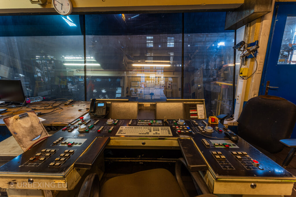 A central control console at the Halo Steelrings / Cockerill Forges & Ringmill factory, depicting a close-up of various switches, levers, and buttons used for managing industrial operations. The backdrop features a large, grimy window providing a view into the dim, deserted factory floor, with reflections of lights and machinery barely visible. The console is flanked by a chair and is accompanied by a telephone, indicating its former role as a pivotal command center for factory operations.