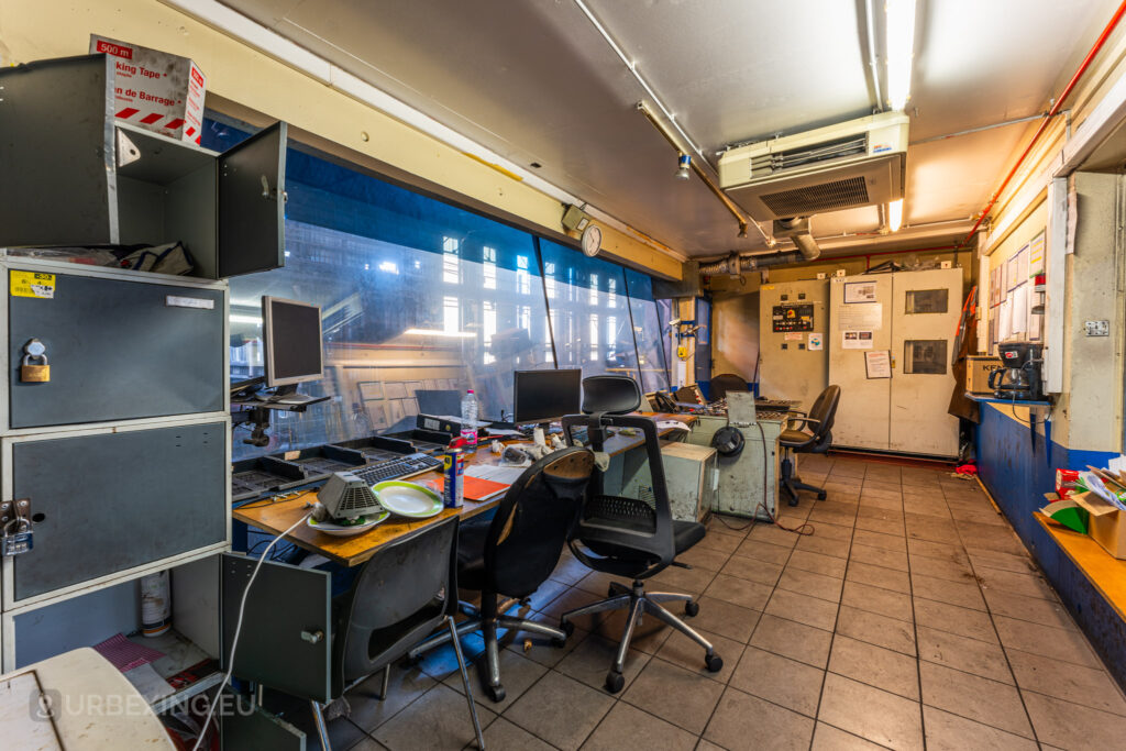 The interior of a control room at the abandoned Halo Steelrings / Cockerill Forges & Ringmill, featuring several old office chairs, dusty computer monitors, and cluttered desks. The room is separated from the factory floor by a large blue-tinted window, and the atmosphere suggests a space left hastily with items scattered around. Various papers, cabinets, and an air conditioning unit contribute to the sense of past functionality in an otherwise abandoned environment.