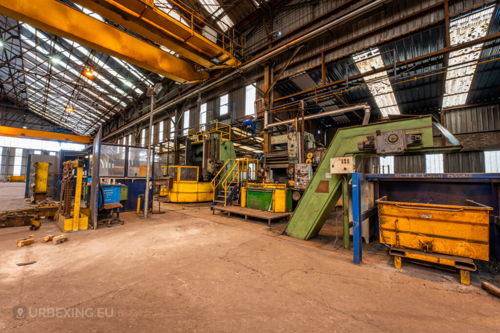 An interior view of the abandoned Halo Steelrings / Cockerill Forges & Ringmill factory, featuring various large industrial machines and equipment scattered across the factory floor. The machinery is painted in bright colors like green and yellow, indicating different functions. Overhead, a massive crane is mounted on beams. The high ceiling is lined with corrugated metal sheets, and sunlight filters through gaps, illuminating the spacious and dusty environment.