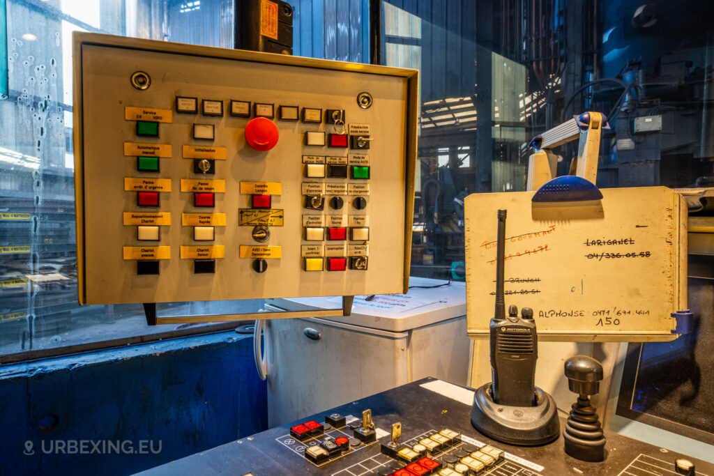 A close-up view of a control panel at Halo Steelrings / Cockerill Forges & Ringmill, showcasing an array of buttons labeled in French, including an emergency stop button prominently displayed in red. The panel, situated next to a board with handwritten notes and a two-way radio, suggests an environment where communication and control were crucial. The background shows a partially visible, dirty factory window, providing a glimpse of the industrial area beyond.