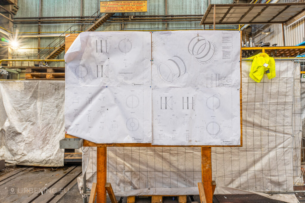 Engineering blueprints pinned to a wooden board in the abandoned Ateliers de La Meuse, Liège, Belgium, with a high-visibility yellow safety vest hanging nearby and industrial staircases and signs in the background.