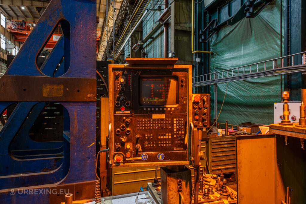 Close-up of an old control panel with a screen displaying numerical data in the abandoned Ateliers de La Meuse, Liège, Belgium, surrounded by metal structures and work tools under warm lighting.