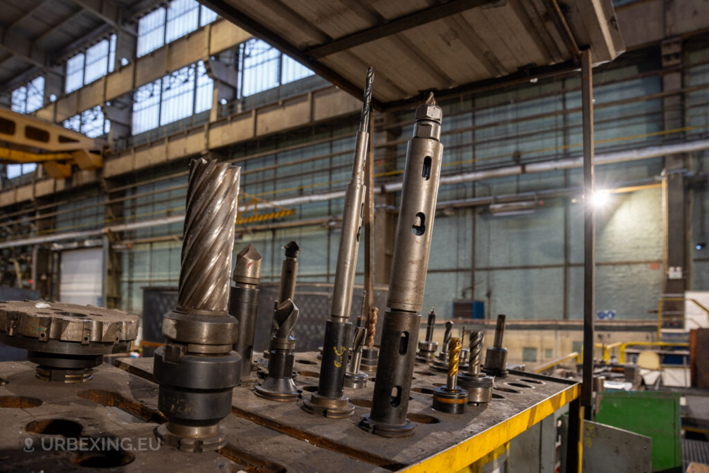 Close-up of various industrial drill bits and cutting tools on a workbench in the abandoned Ateliers de La Meuse, Liège, Belgium, with the large warehouse windows and structural beams visible in the background.