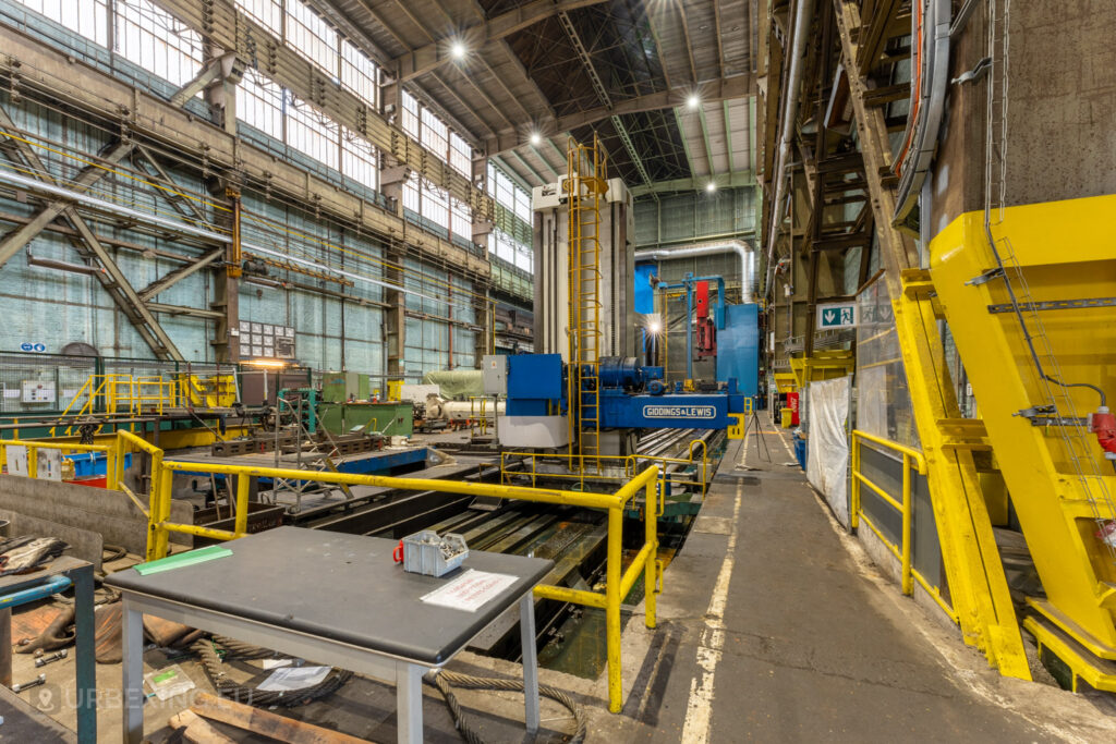 Abandoned industrial workspace at Ateliers de La Meuse in Liège, Belgium, featuring a large blue Giddings & Lewis milling machine, surrounded by yellow safety railings and scattered equipment in a high-ceilinged hall.