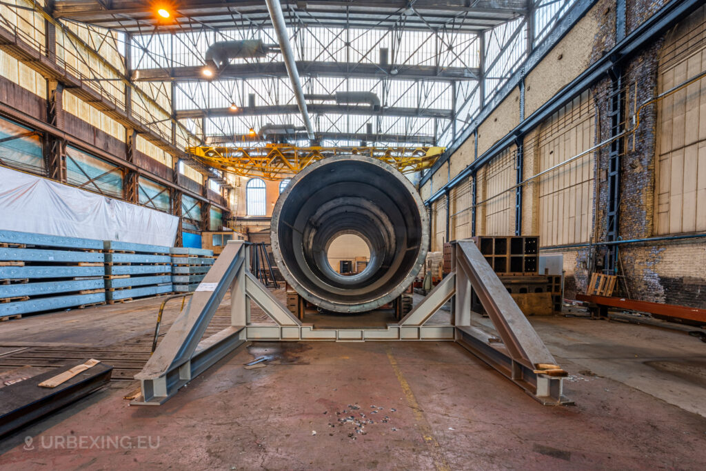 Large cylindrical industrial component inside the abandoned Ateliers de La Meuse in Liège, Belgium, surrounded by steel beams and illuminated by overhead lights in a cavernous, decaying warehouse.