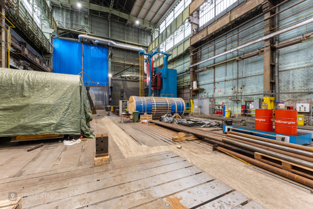 Spacious industrial hall in the abandoned Ateliers de La Meuse in Liège, Belgium, featuring a large blue machine, wooden reels, and oil drums, surrounded by scattered tools and industrial debris.