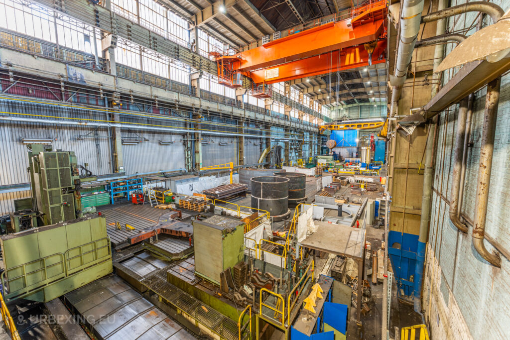 Expansive industrial hall of the abandoned Ateliers de La Meuse in Liège, Belgium, showcasing large cylindrical structures, scattered equipment, and an orange overhead crane dominating the space.