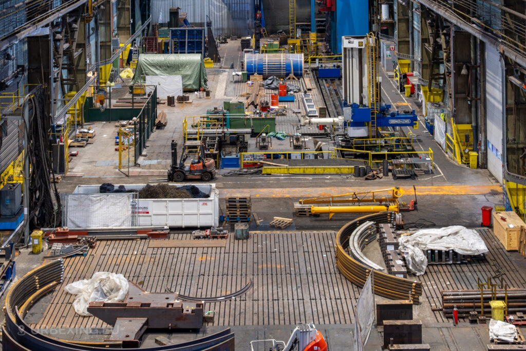 High-angle view of the abandoned industrial hall at Ateliers de La Meuse in Liège, Belgium, with scattered heavy machinery, a forklift, metal components, and a blue Giddings & Lewis milling machine surrounded by yellow safety barriers.