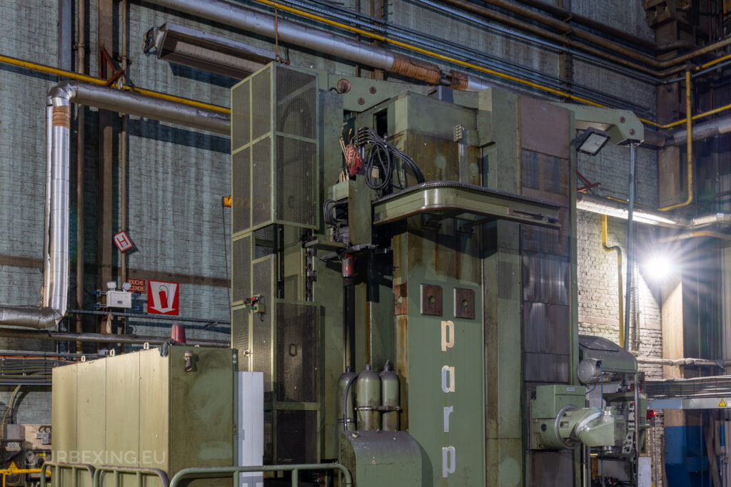 Close-up of a large green industrial machine labeled ‘parp’ in the abandoned Ateliers de La Meuse, Liège, Belgium, with surrounding pipes, cables, and wall-mounted safety signage.