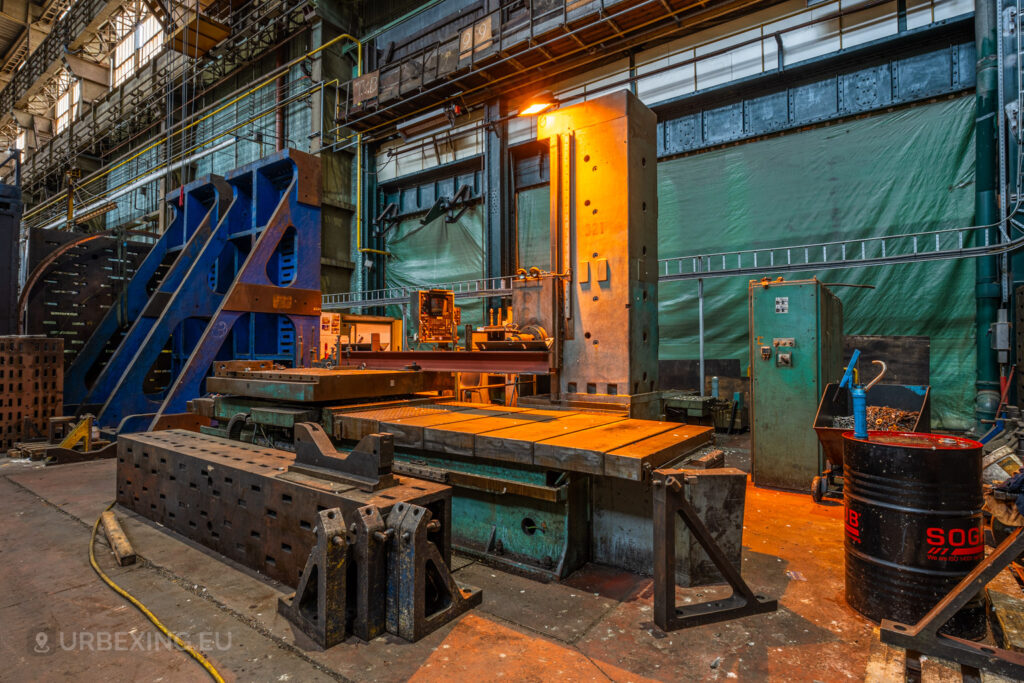 Industrial machinery setup in the abandoned Ateliers de La Meuse, Liège, Belgium, featuring a large metal workbench, blue structural beams, and equipment illuminated by a single orange light in a dimly lit workshop.