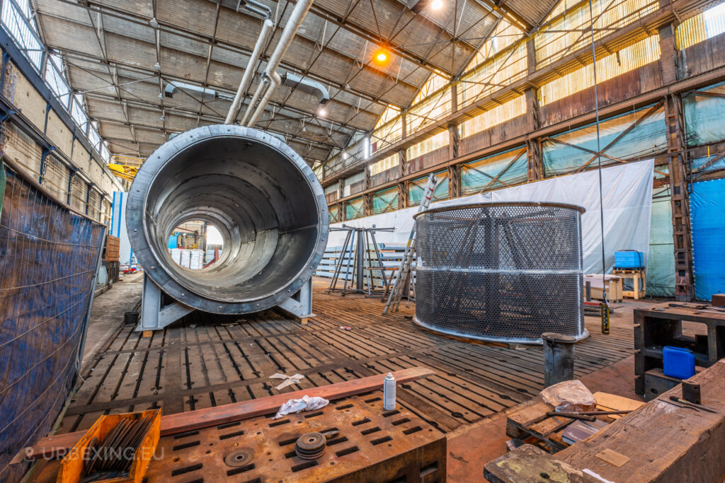 Massive metal tube and perforated steel structure inside the abandoned Ateliers de La Meuse in Liège, Belgium, surrounded by scattered tools and wooden planks in a cavernous, sunlit warehouse.