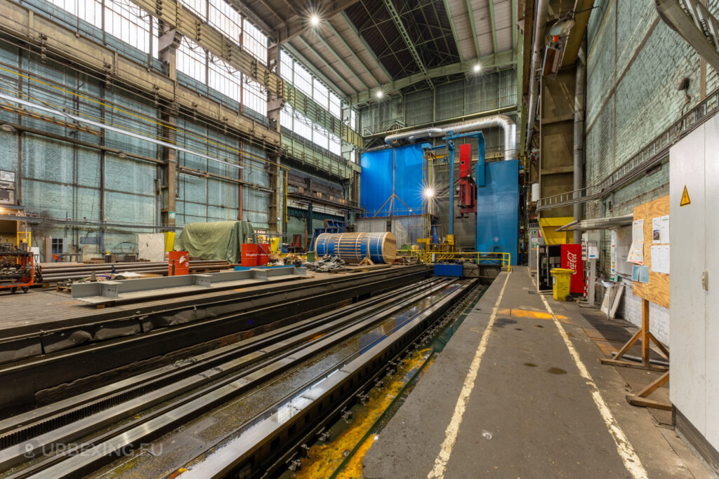 Large industrial workshop inside the abandoned Ateliers de La Meuse in Liège, Belgium, featuring tracks, heavy machinery, and various metal components scattered across the expansive, high-ceilinged space.