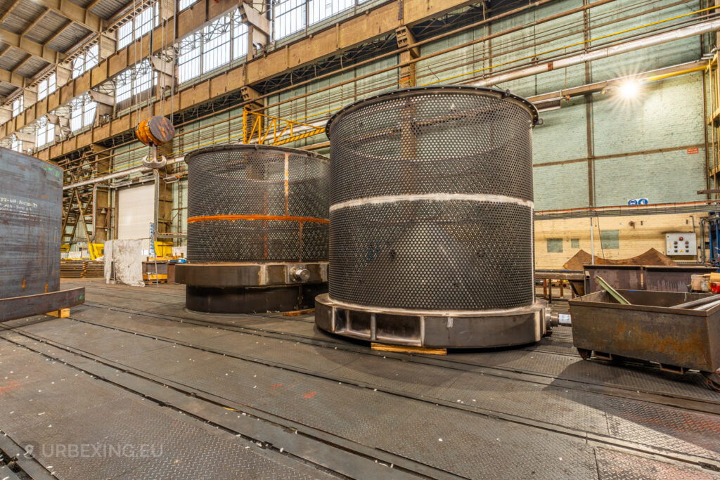 Massive cylindrical metal structures with perforated surfaces stand on a steel platform in the abandoned Ateliers de La Meuse, Liège, Belgium, with industrial cranes and large windows in the background.