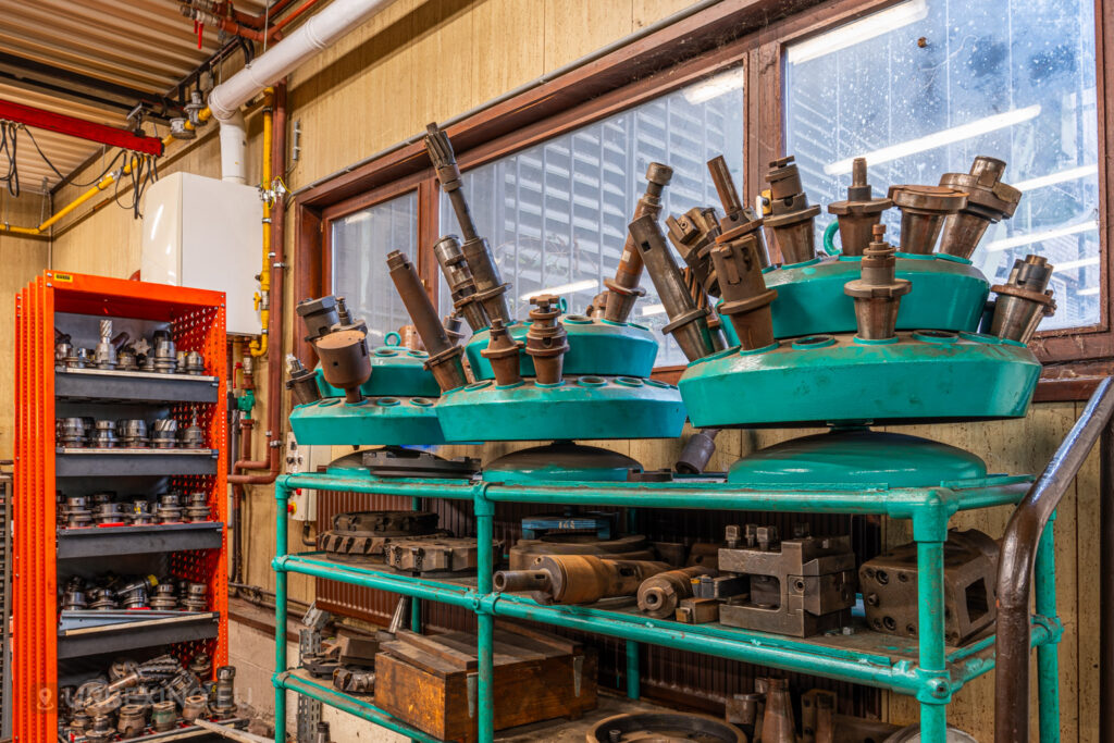 Close-up of industrial metal tool heads and components stored on green metal shelves in the abandoned Ateliers de La Meuse, Liège, Belgium, with an orange storage rack in the background and dusty windows.