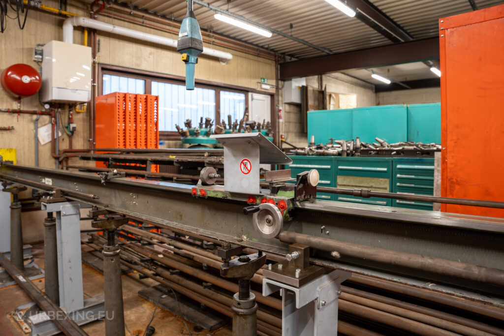 Close-up of a metalworking station with a rail-mounted industrial tool and hanging blue inspection lamp in the abandoned Ateliers de La Meuse, Liège, Belgium. The workshop features teal and orange cabinets and various scattered metal rods.