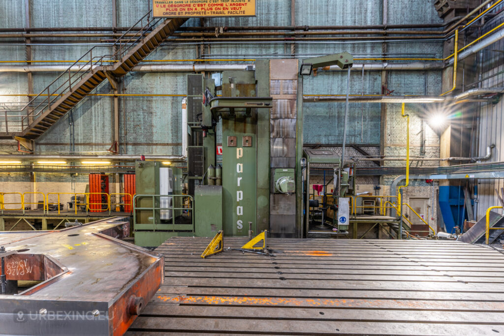 Large industrial machine labeled ‘parpas’ in the abandoned Ateliers de La Meuse, Liège, Belgium, with a massive metal work platform in the foreground, staircases, and overhead pipes lining the walls.