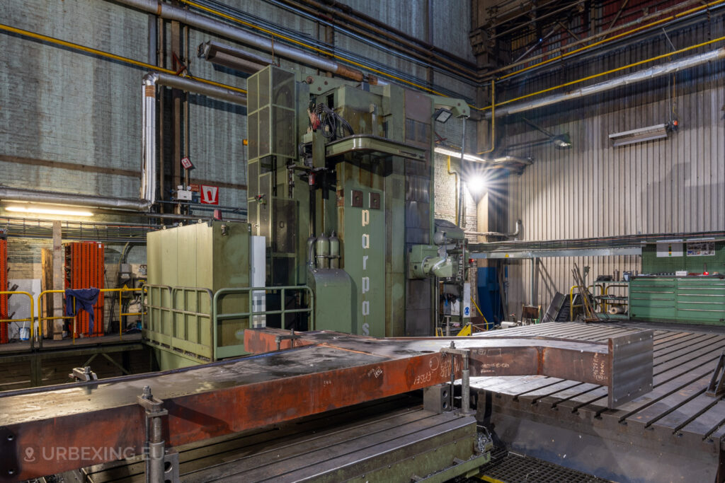 Large green industrial machine labeled ‘parpas’ in the abandoned Ateliers de La Meuse, Liège, Belgium, with rusted metal components on a heavy-duty workbench and surrounding pipes and cables.