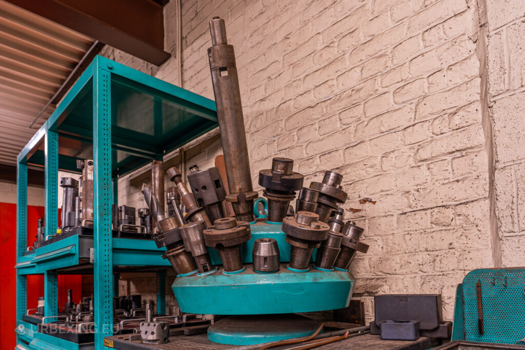 Collection of large metal tool heads and industrial equipment stored on teal metal shelves in the abandoned Ateliers de La Meuse, Liège, Belgium, with a textured white brick wall in the background.