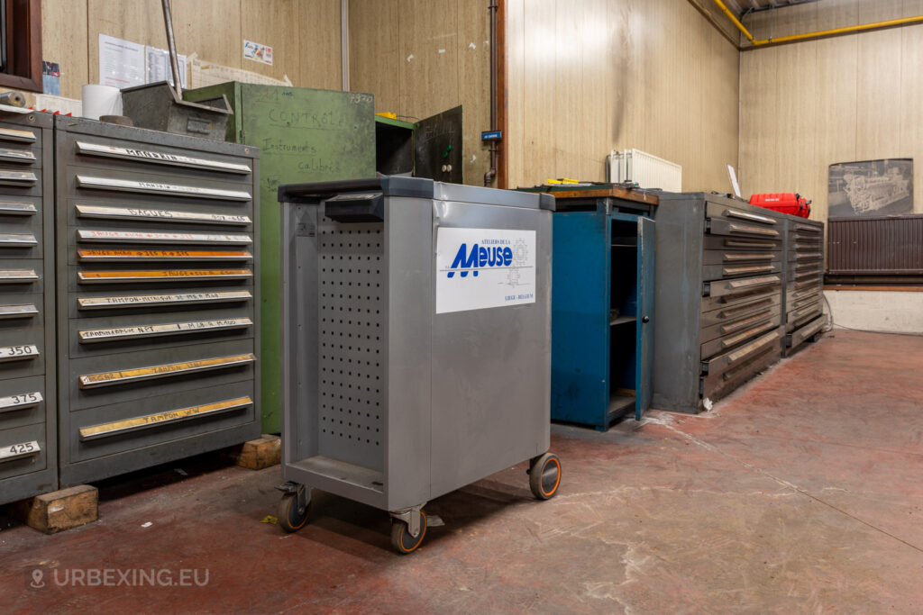 Metal tool cabinets and a wheeled cart labeled ‘Ateliers de La Meuse’ in an abandoned workshop in Liège, Belgium, with worn flooring and a wall decorated with old industrial images.