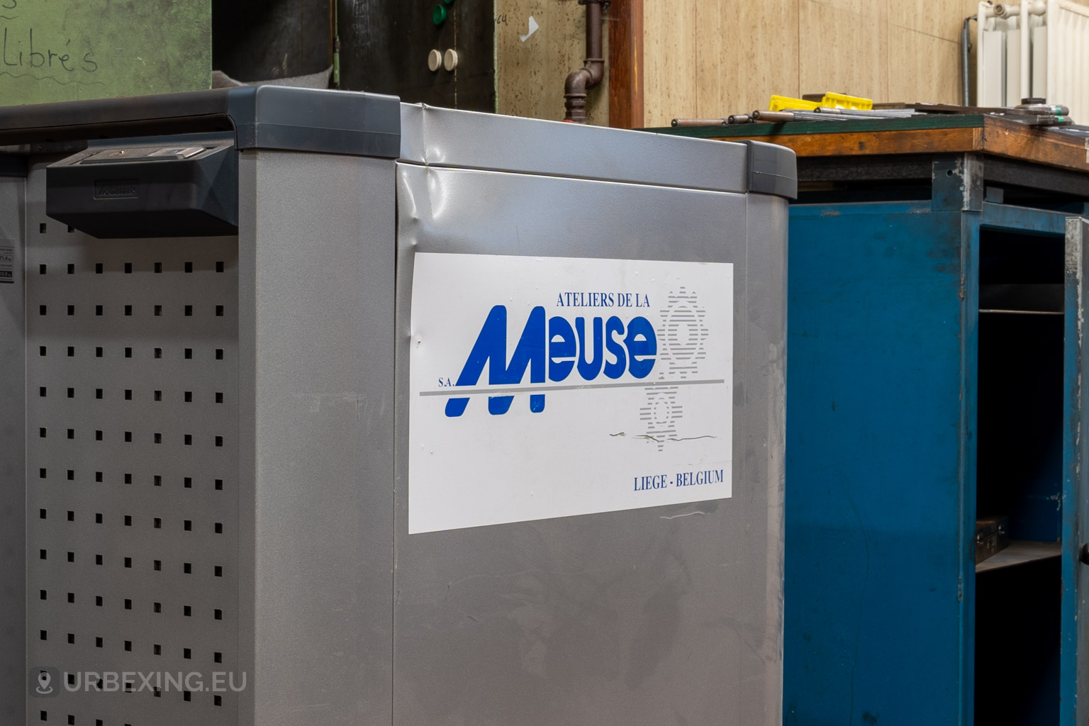 Close-up of a dented metal tool cabinet featuring the Ateliers de La Meuse logo in blue text, Liège, Belgium, with scattered workshop tools in the background.