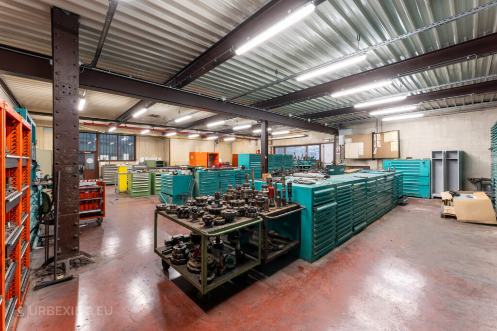 Abandoned workshop room at Ateliers de La Meuse in Liège, Belgium, filled with green tool cabinets, orange shelving units, and carts holding various metal components, illuminated by fluorescent lighting.