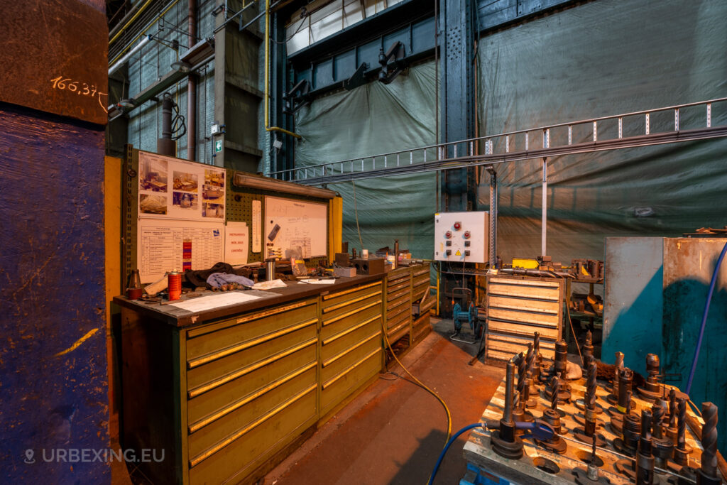 Workstation in the abandoned Ateliers de La Meuse, Liège, Belgium, featuring green tool cabinets, a cluttered workbench with notes and drawings, and a pallet of industrial drill bits under warm lighting.