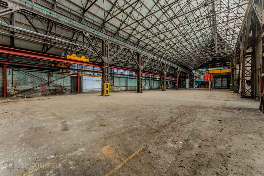 Vast abandoned industrial hall with steel trusses and remnants of machinery at the ArcelorMittal Marchin plant in Liège, Belgium.