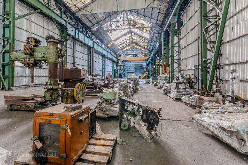 Decaying machinery and industrial equipment rest untouched in the abandoned ArcelorMittal Marchin electro-galvanization plant in Liège, Belgium. The high glass-paneled ceiling and green steel girders emphasize the vastness of the now-silent hall, once bustling with industrial activity.