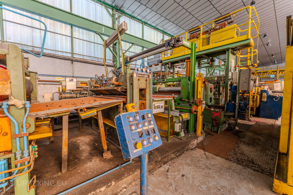 Abandoned industrial machinery with a blue control panel and vibrant green and yellow components at the ArcelorMittal Marchin plant in Liège, Belgium.