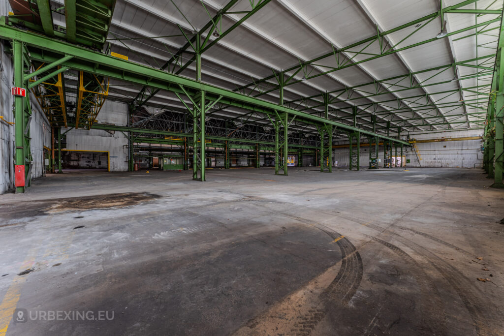 Empty industrial hall with green steel beams and tire marks on the concrete floor at the abandoned ArcelorMittal Marchin plant in Liège, Belgium.