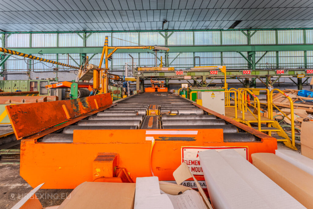 Orange industrial conveyor system with rollers and control stations inside the abandoned ArcelorMittal Marchin plant in Liège, Belgium.