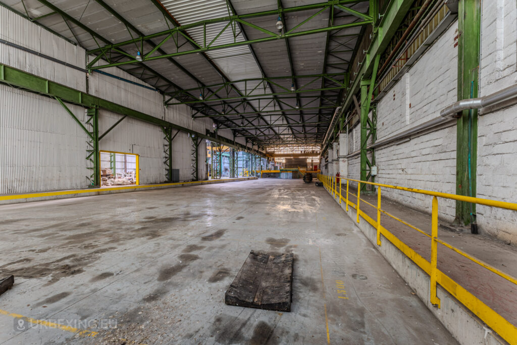 The industrial corridor of the abandoned ArcelorMittal Marchin electro-galvanization plant in Liège, Belgium, highlights its desolate charm. Green steel beams and bright yellow safety railings contrast with the muted concrete flooring, reflecting the site’s former functionality and its current state of urban decay.