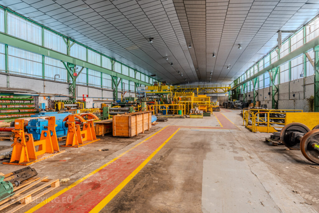 Spacious industrial hall with abandoned machinery, vibrant orange and blue equipment, and organized storage at the ArcelorMittal Marchin plant in Liège, Belgium.