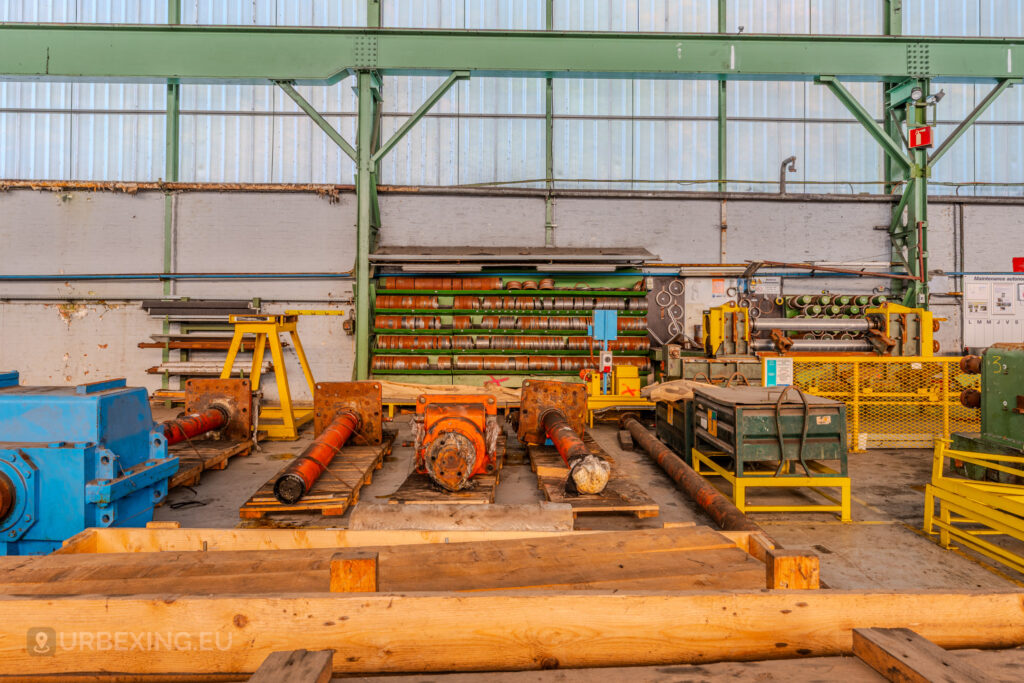 Stored industrial parts, including orange rollers and blue machinery, at the abandoned ArcelorMittal Marchin plant in Liège, Belgium.