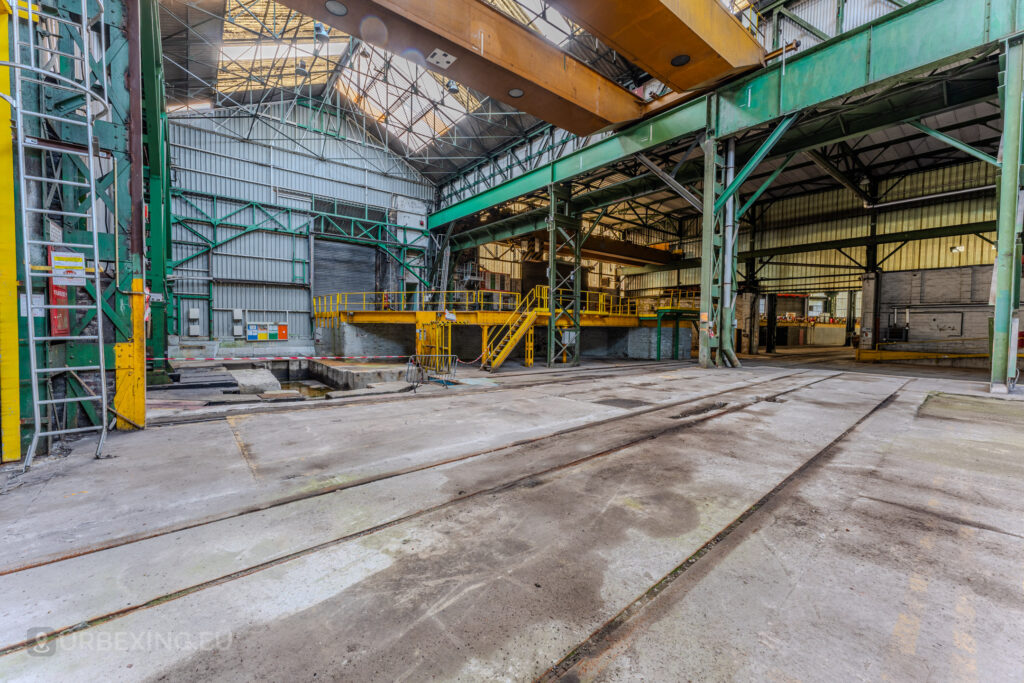 Abandoned industrial workshop with rail tracks, yellow safety platforms, and green steel beams at the ArcelorMittal Marchin plant in Liège, Belgium.