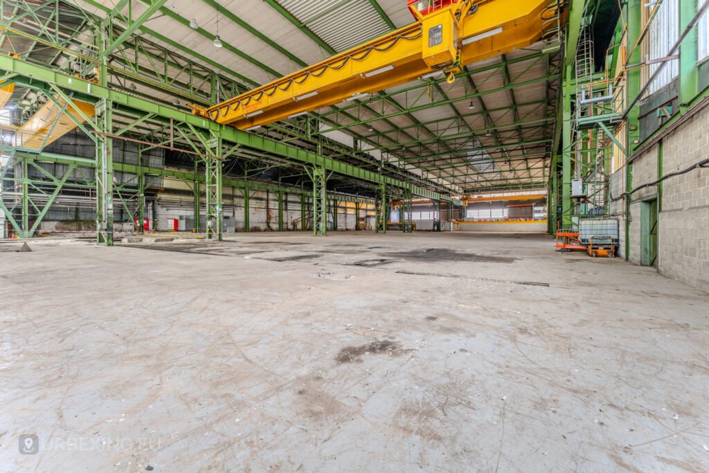 Inside the abandoned ArcelorMittal Marchin electro-galvanization plant in Liège, Belgium. The expansive industrial hall, now deserted, showcases towering green steel girders and a bright yellow overhead crane, evoking the site’s past as a hub of manufacturing before its closure in 2013.