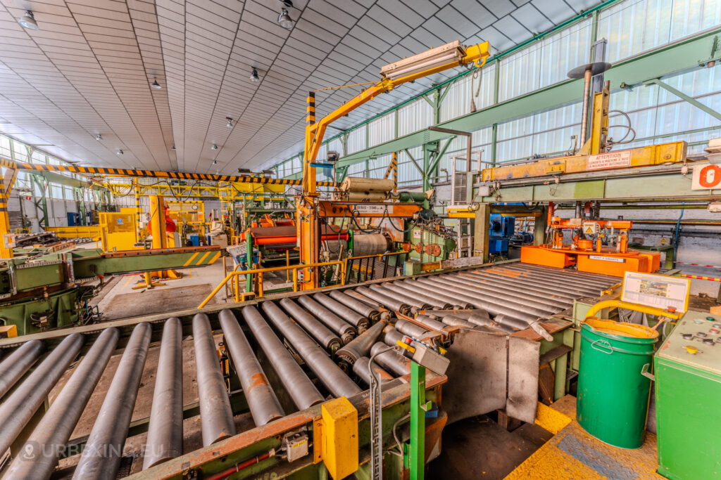 Industrial roller conveyor and vibrant machinery inside the abandoned ArcelorMittal Marchin plant in Liège, Belgium, featuring yellow and orange equipment.