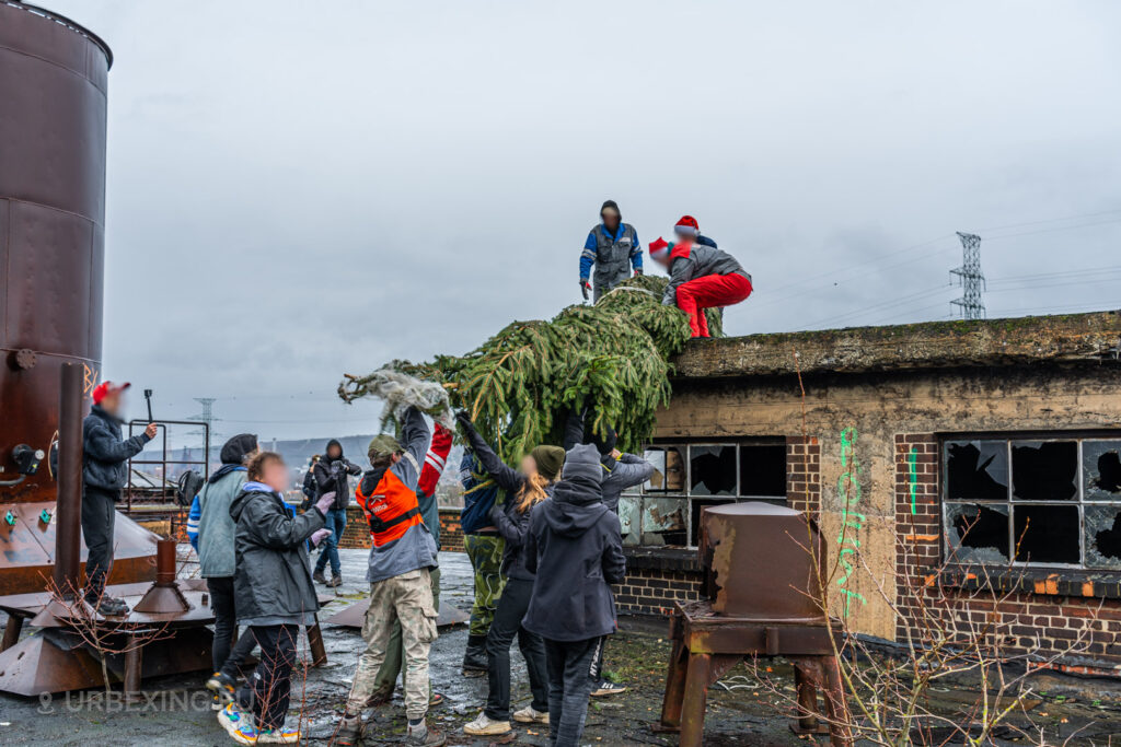 Urban explorers collaborate to hoist a Christmas tree onto the rooftop of the abandoned HF6 power plant in Seraing, Belgium, blending festive cheer with the rugged charm of urban exploration.