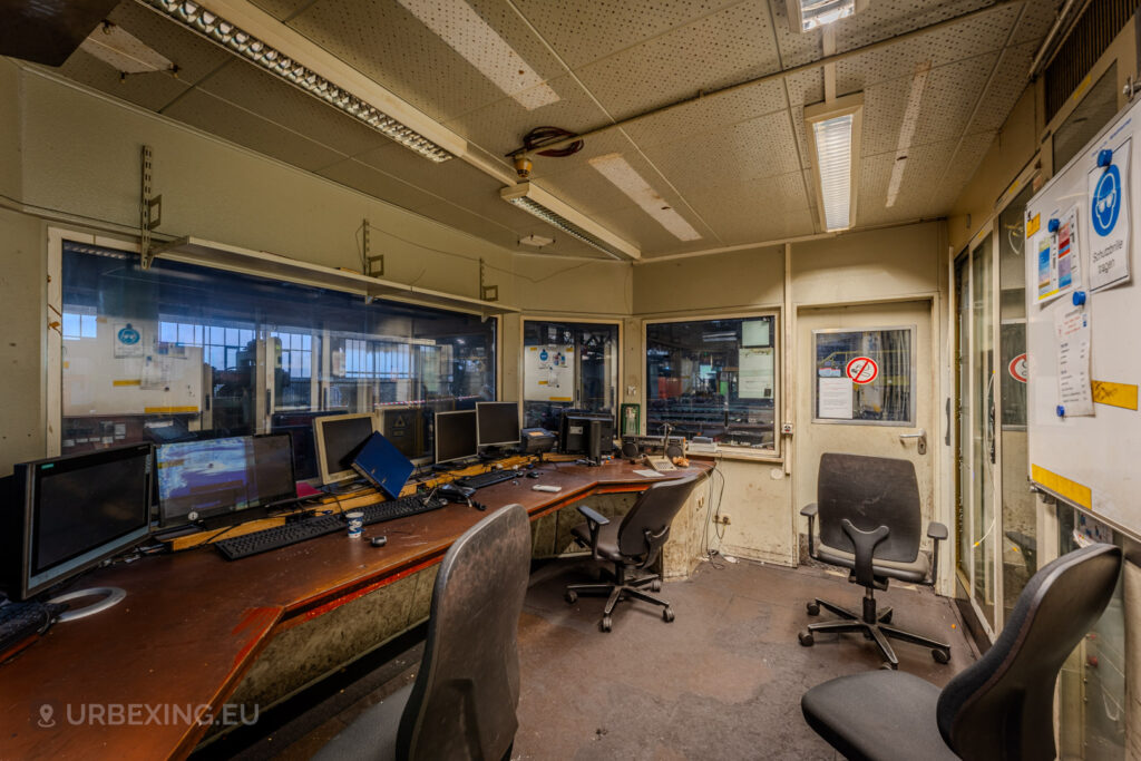 Control room in the abandoned Vallourec steel mill in Düsseldorf-Rath, with dusty computer monitors, swivel chairs, and glass windows overlooking the industrial floor, evoking the end of an era in steel manufacturing.”