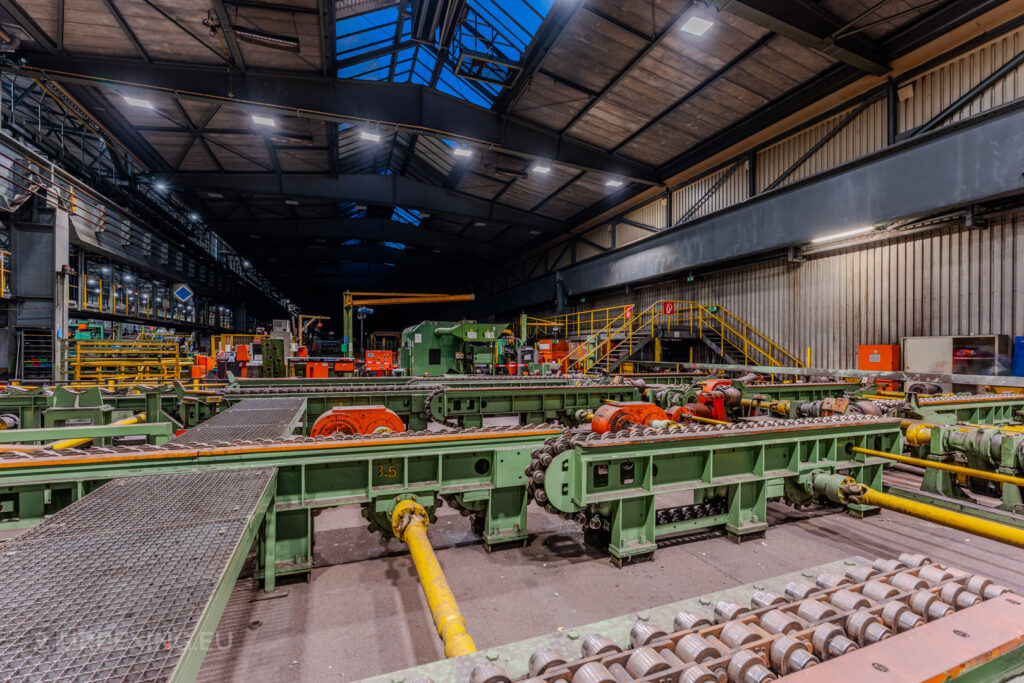 Detailed view of conveyor systems and machinery in the abandoned Vallourec steel mill in Düsseldorf-Rath, featuring green and orange industrial components under a skylit arched ceiling.